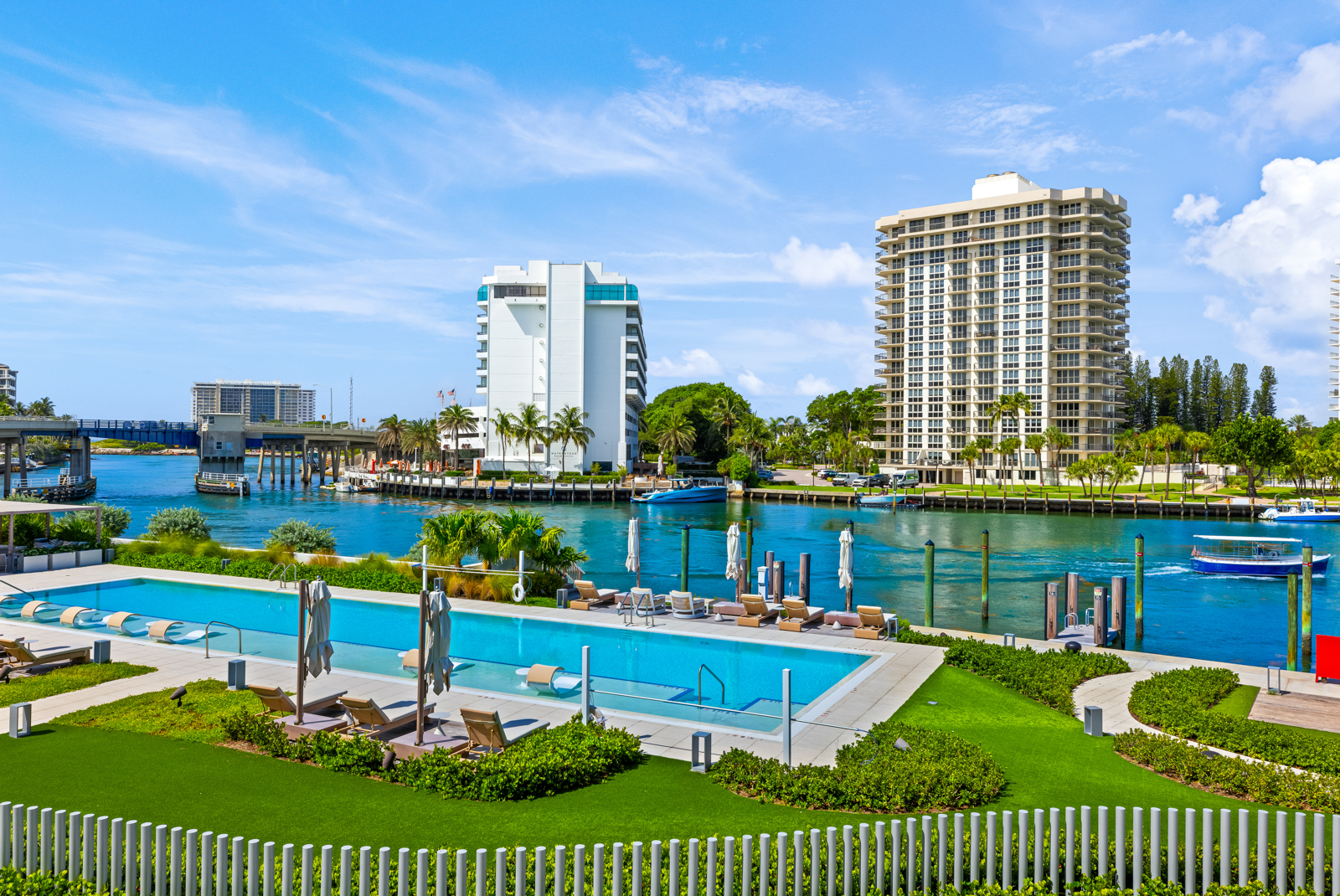 This image showcases a luxurious outdoor pool area with modern lounge chairs and umbrellas, set against a backdrop of waterfront views and high-rise buildings. The manicured green lawn and white picket fence add to the upscale ambiance, creating an inviting and serene environment. The scene is captured from a high-angle perspective, emphasizing the pool's design and the surrounding landscape.