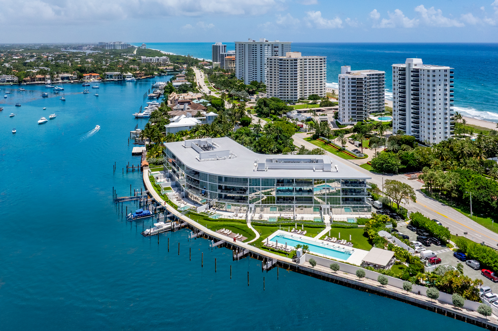 This aerial shot showcases a luxurious waterfront property featuring a modern, curved building with expansive glass windows and a flat roof. The property includes a swimming pool, manicured lawns, and a private dock with boats. The surrounding area features high-rise condominiums, lush greenery, and the ocean, creating an impression of upscale coastal living.