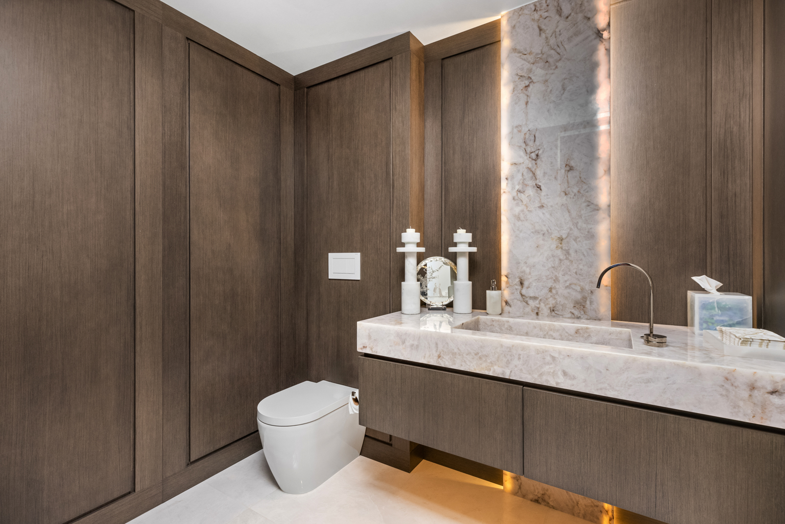 This is a modern guest bathroom featuring a floating vanity with a marble countertop and integrated sink. The walls are clad in dark wood paneling, complemented by a marble backsplash with integrated lighting. A sleek, wall-mounted toilet adds to the minimalist aesthetic, creating a sophisticated and clean space.