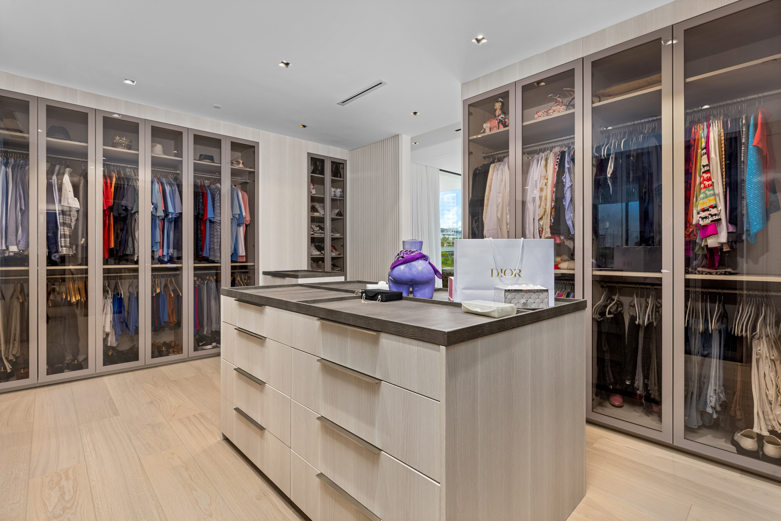 This is a spacious walk-in closet featuring custom cabinetry with glass doors, providing ample storage for clothing and accessories. A central island with drawers offers additional surface space and organization. The light wood flooring and neutral color palette create a bright and luxurious atmosphere, enhancing the appeal for potential buyers.