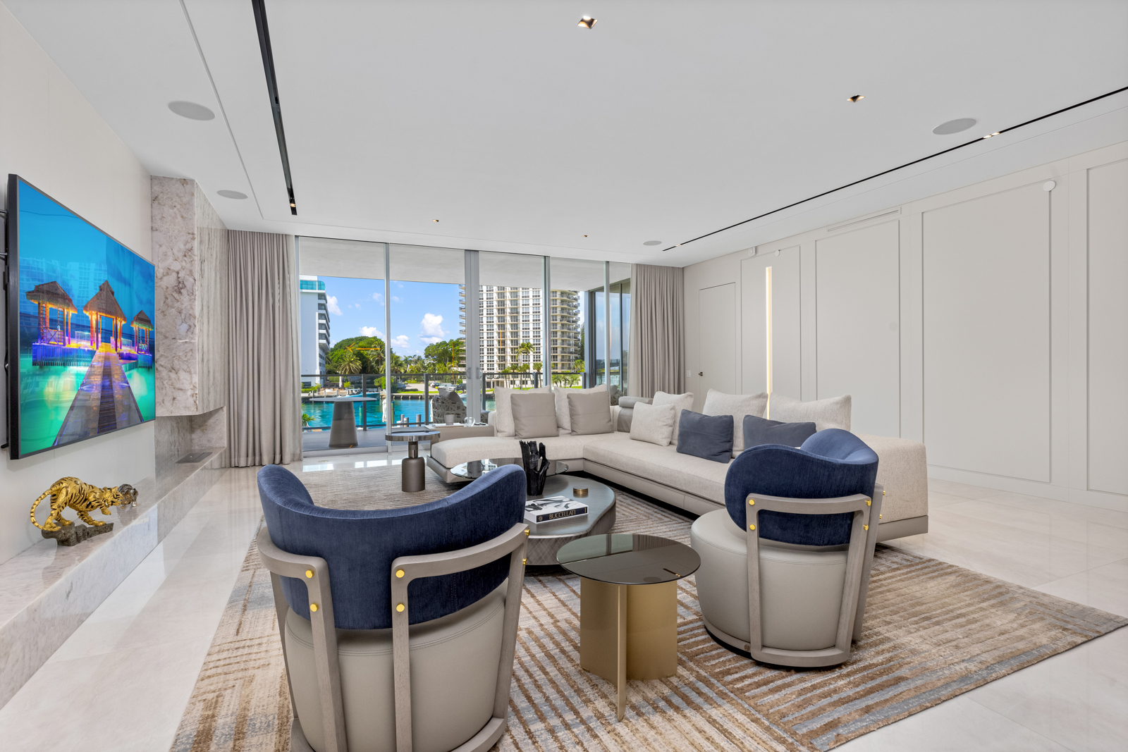 This is a luxurious living room featuring a large sectional sofa with throw pillows, two stylish armchairs, and a modern coffee table. The room has floor-to-ceiling windows offering a view of the water, and a large television is mounted on a marble accent wall. The overall design is contemporary and elegant, creating a comfortable and sophisticated living space.