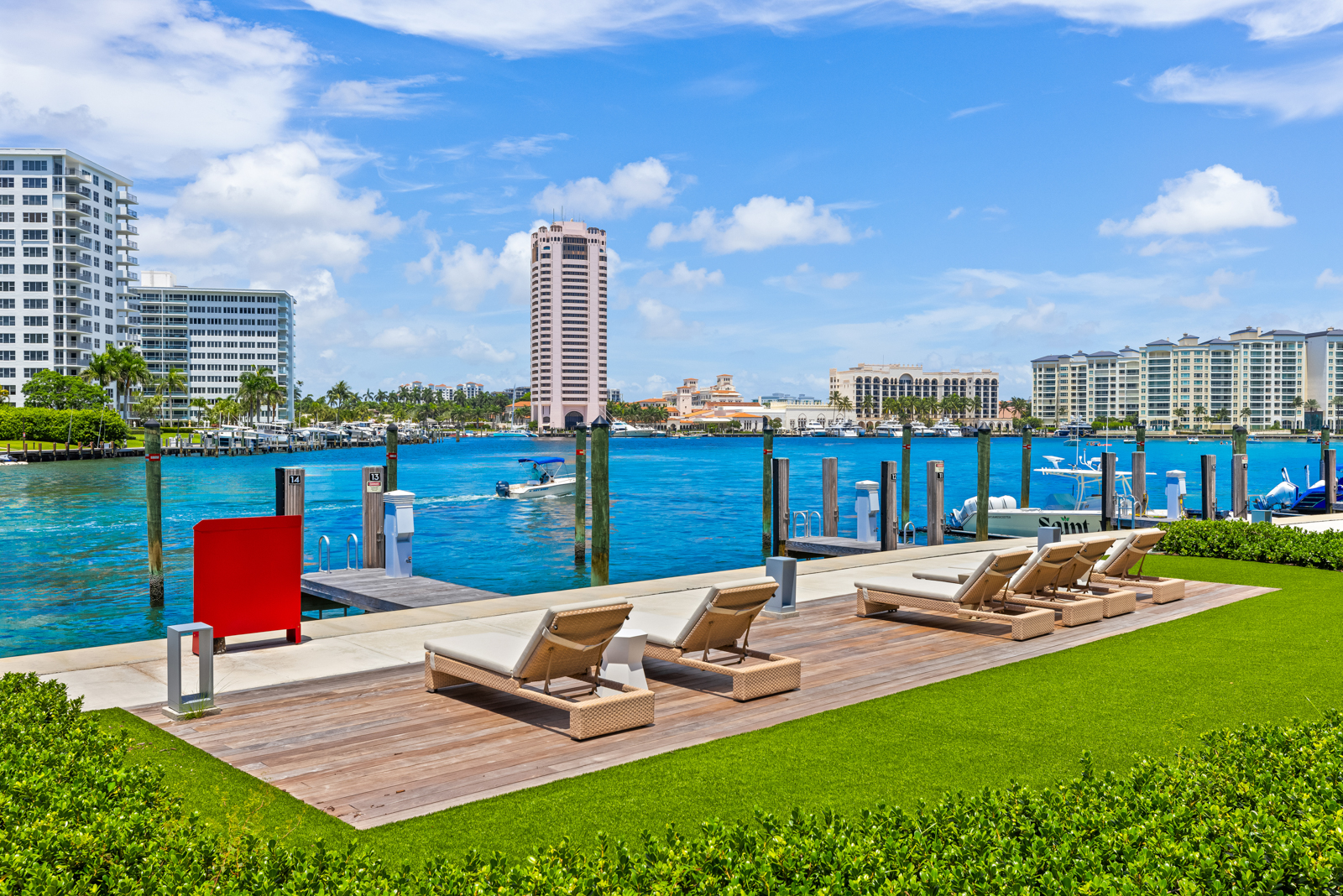 This image showcases a luxurious waterfront patio or deck area, featuring multiple lounge chairs arranged on a wooden deck adjacent to a manicured lawn. The scene offers a stunning view of the waterway, with modern buildings and boats visible in the background, creating an inviting and upscale outdoor living space. The perspective is from a low angle, emphasizing the depth and breadth of the waterfront view.