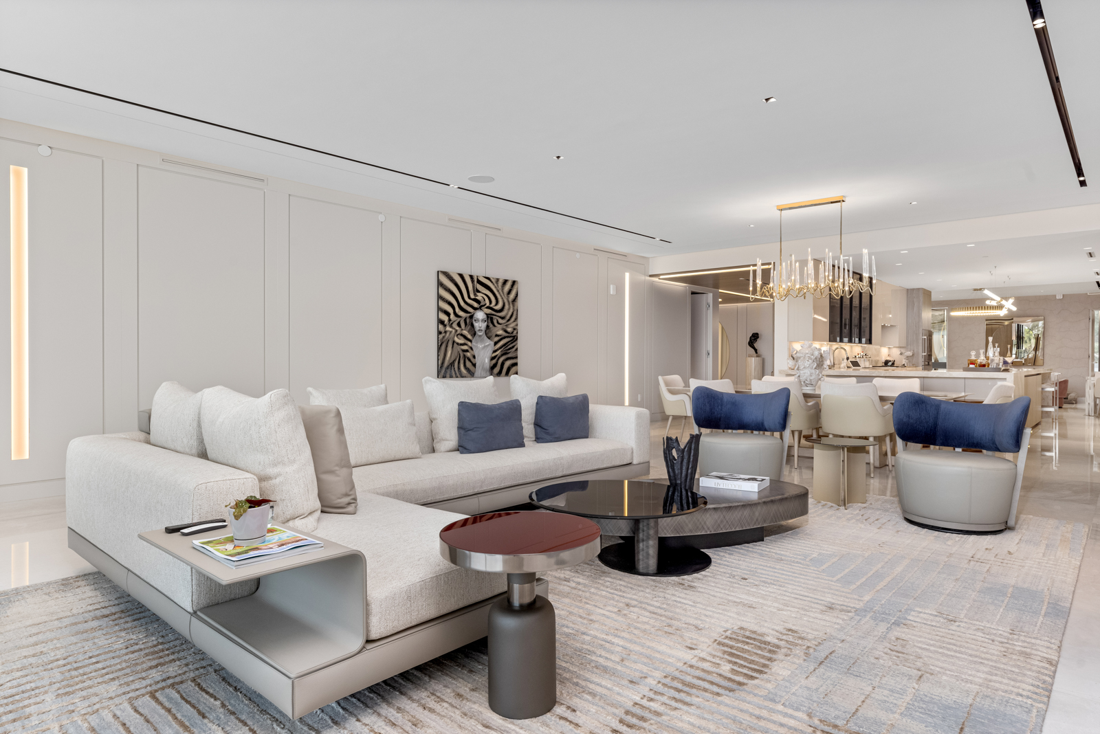 This is a bright and elegant living room featuring a large sectional sofa with throw pillows, a modern coffee table arrangement, and stylish accent chairs. The room is decorated in neutral tones with pops of blue, complemented by a patterned area rug and contemporary artwork. The open floor plan seamlessly connects to the dining area, enhancing the sense of spaciousness and luxury.