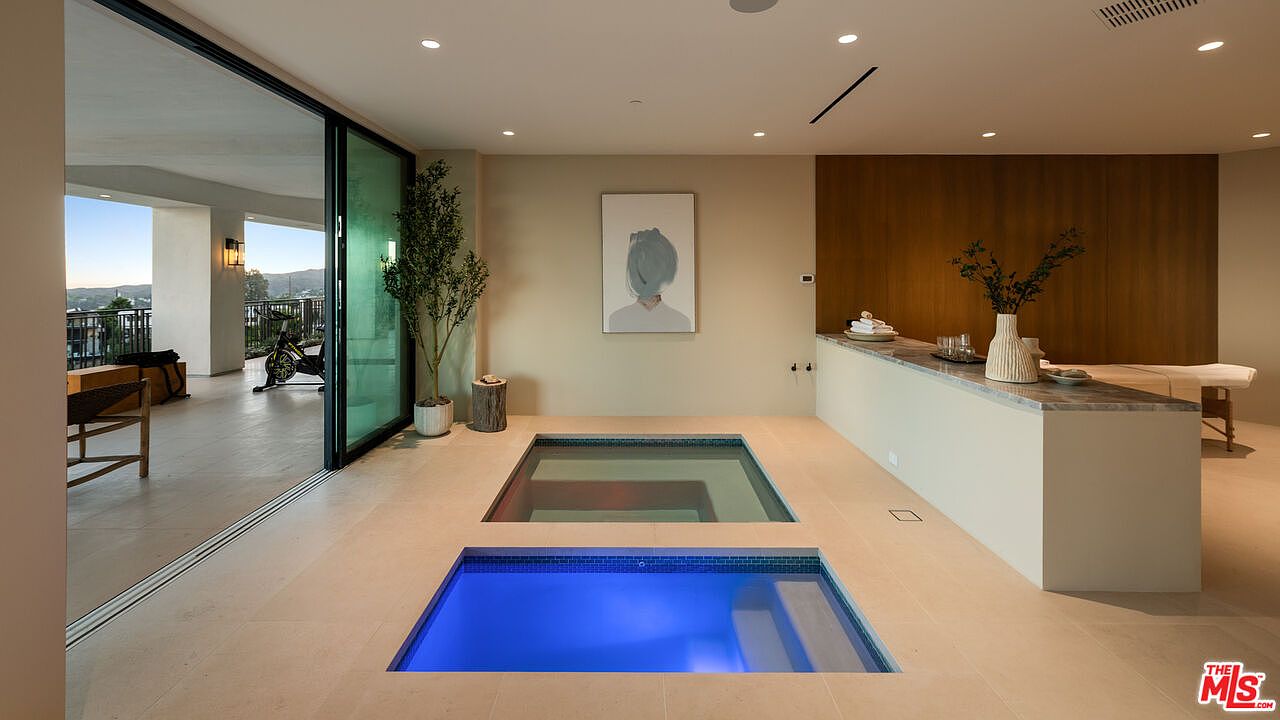 This luxurious indoor spa and wellness area features a sunken pool with blue lighting and an adjacent dry area, seamlessly connected to an outdoor balcony through large sliding glass doors. The space includes a sleek stone-topped counter, a potted plant, and minimalist wall art, creating a serene and high-end atmosphere. The perspective is wide and centered, highlighting the transition between the indoor relaxation zone and the exterior terrace.