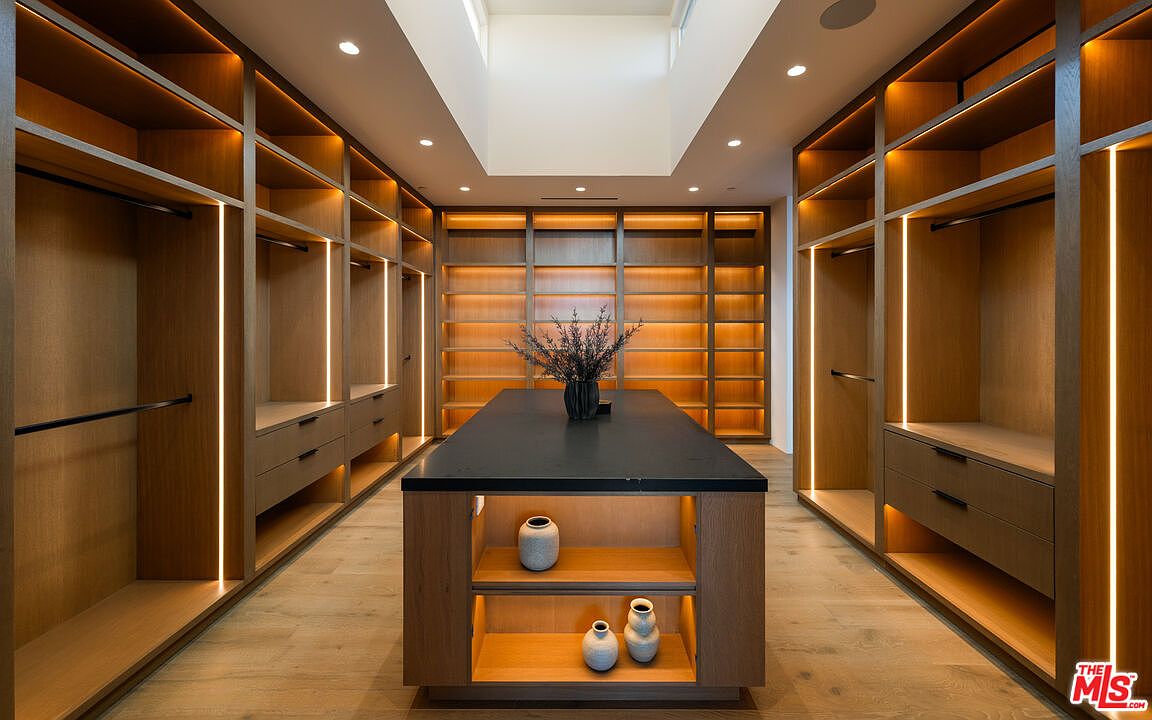 This luxurious walk-in closet features extensive custom light wood cabinetry with integrated vertical LED lighting throughout. A central island with a dark countertop provides additional storage and display space, while a skylight above fills the room with natural light. The space is designed with a clean, modern aesthetic, emphasizing organization and high-end finishes.