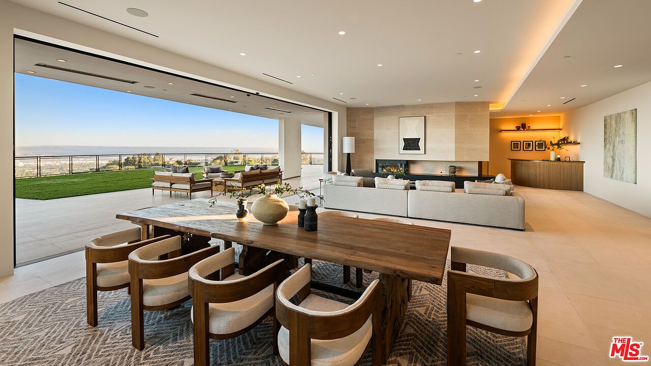 This expansive, open-concept living and dining area features a seamless transition to an outdoor patio through massive, retractable glass walls that frame a panoramic view. The space is anchored by a large, light-toned sectional sofa facing a stone-clad chimney breast with a built-in fireplace, while a substantial wooden dining table sits in the foreground. The interior design emphasizes a neutral color palette, clean lines, and a sophisticated, airy atmosphere suitable for luxury entertaining.