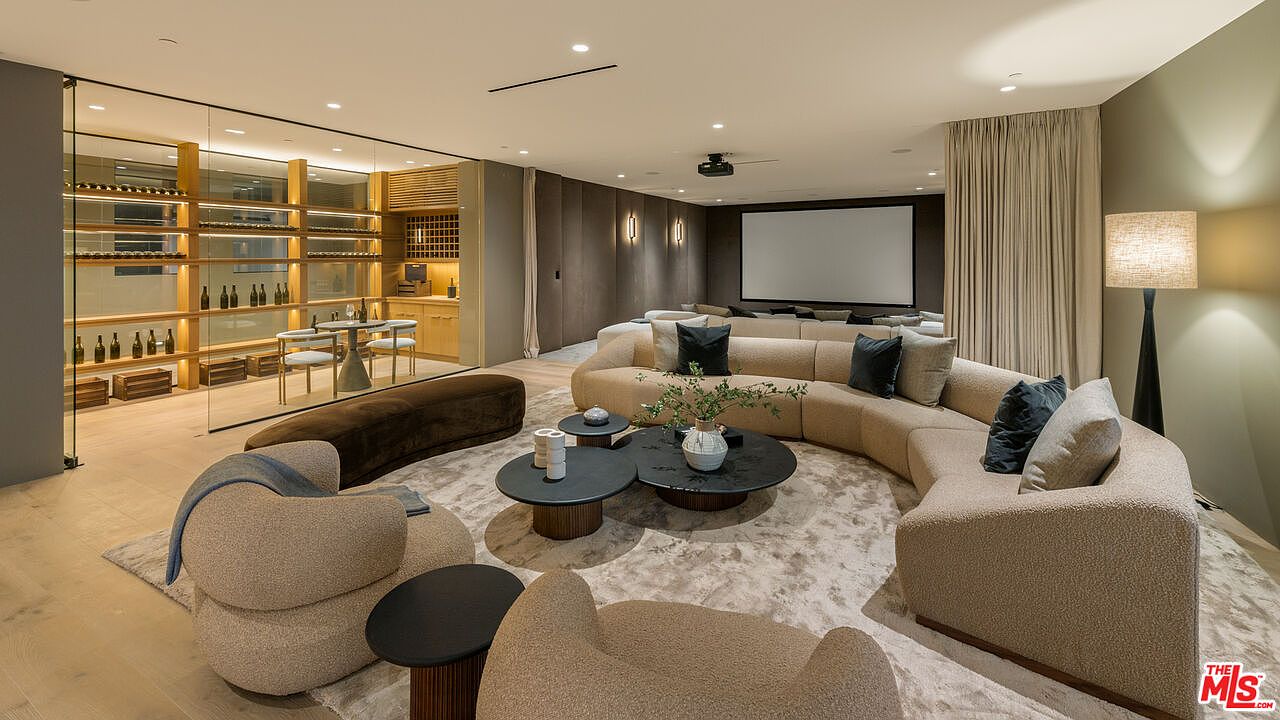 This sophisticated media and lounge area features a large, curved sectional sofa arranged around a circular coffee table on a textured area rug. To the left, a glass-enclosed wine display adds a luxurious touch, while the far wall houses a projection screen for home theater viewing. The room is illuminated by recessed lighting and a tall floor lamp, creating a warm, inviting atmosphere perfect for relaxation or entertainment.