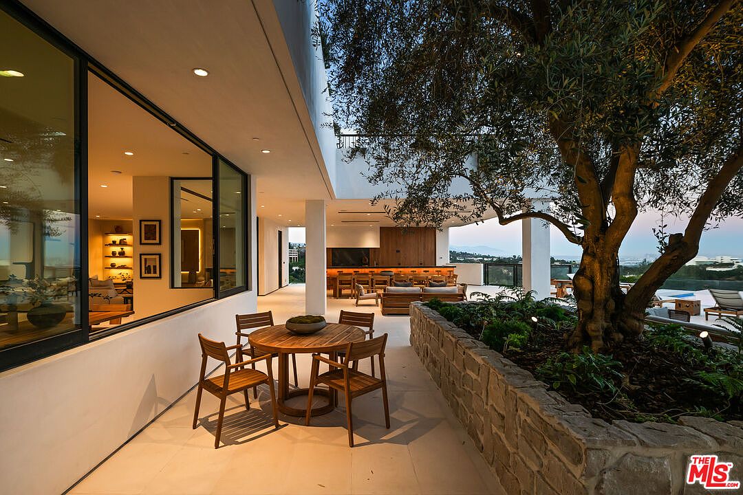 This expansive, modern outdoor patio features a dining area with a circular wooden table and chairs set against a backdrop of large windows looking into the home's interior. A stone-walled planter box houses a mature olive tree, adding a natural element to the sleek, contemporary architecture. In the background, an outdoor kitchen and lounge area overlook a scenic view, creating a sophisticated space for alfresco entertaining.
