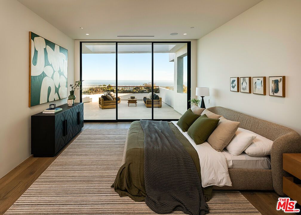 This spacious primary bedroom features a contemporary design with a neutral color palette and an emphasis on indoor-outdoor living. A large bed with olive-toned bedding sits on a striped area rug, facing floor-to-ceiling glass sliding doors that open onto a private balcony with ocean views. The room is accented by a dark wood sideboard, abstract wall art, and a series of framed prints, creating a sophisticated and serene atmosphere.