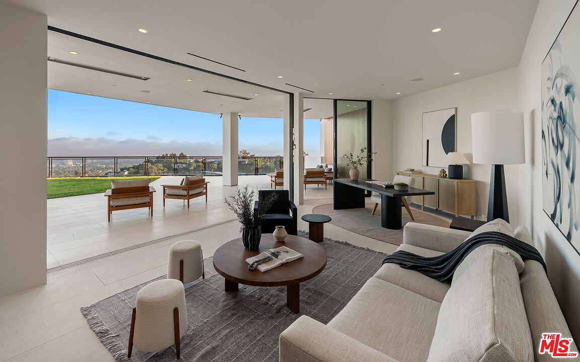 This sophisticated open-concept living area seamlessly transitions to a spacious outdoor patio through expansive glass walls, offering panoramic views of the landscape. The interior features a neutral color palette with a textured area rug, a round wooden coffee table, and contemporary seating, while an adjacent office space provides a functional work area. The design emphasizes a bright, airy atmosphere with recessed lighting and a minimalist aesthetic that highlights the connection between indoor and outdoor living.