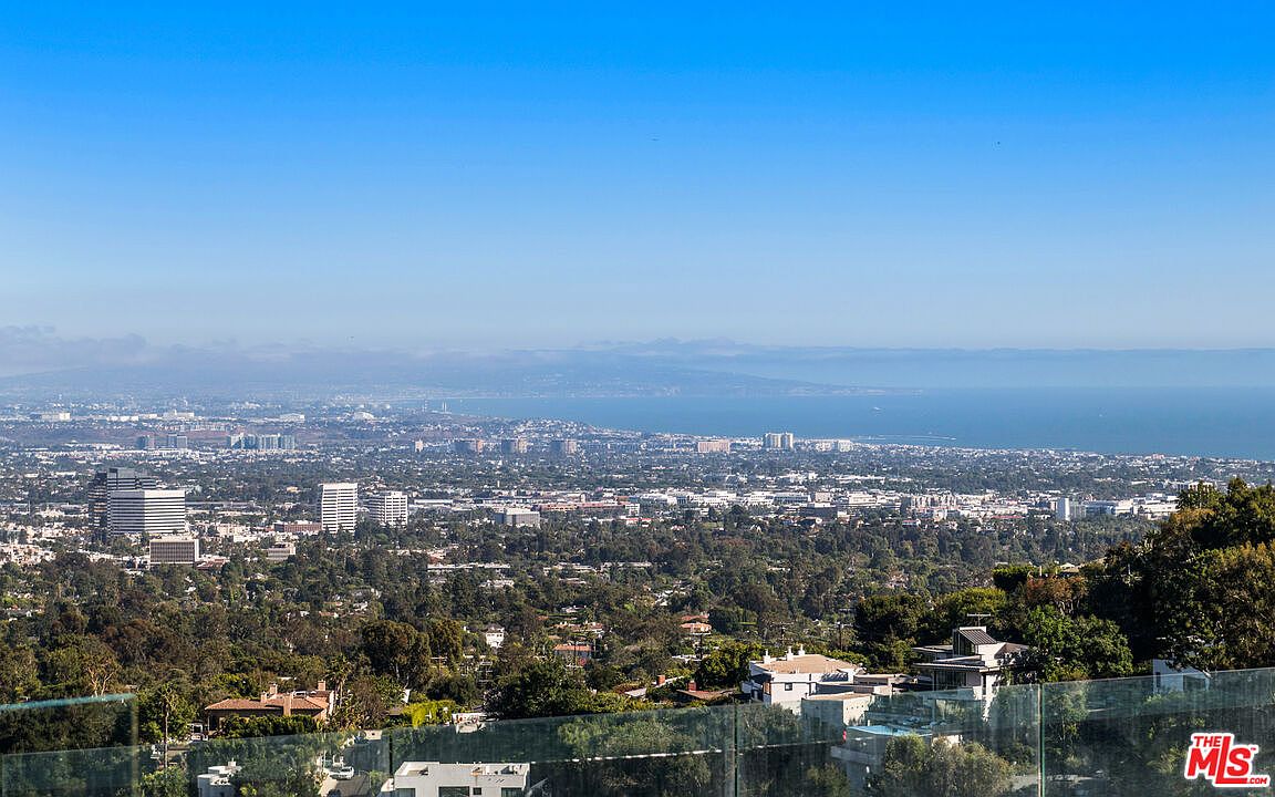 This image captures a sweeping, elevated panoramic view of a sprawling city landscape extending toward the distant coastline and ocean. In the foreground, a glass railing defines the edge of a balcony, providing an unobstructed vantage point over the lush, tree-filled residential neighborhood below. The clear blue sky and bright daylight emphasize the expansive scale and prime location of the property.