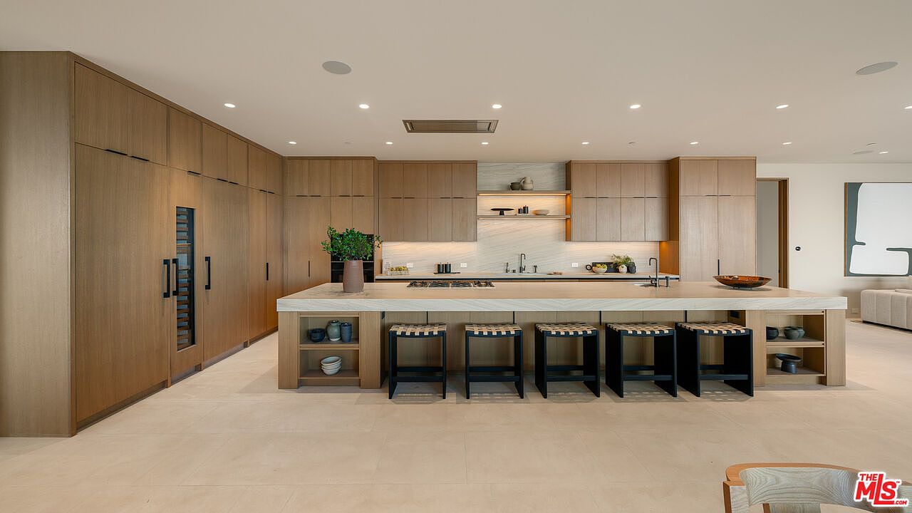 This expansive, modern kitchen features light wood cabinetry and a massive central island with integrated seating for five. The space is characterized by clean lines, a neutral color palette, and a minimalist aesthetic, with a stone backsplash and recessed lighting throughout. The perspective is a wide, eye-level shot that captures the full breadth of the cabinetry and the functional layout of the workspace.