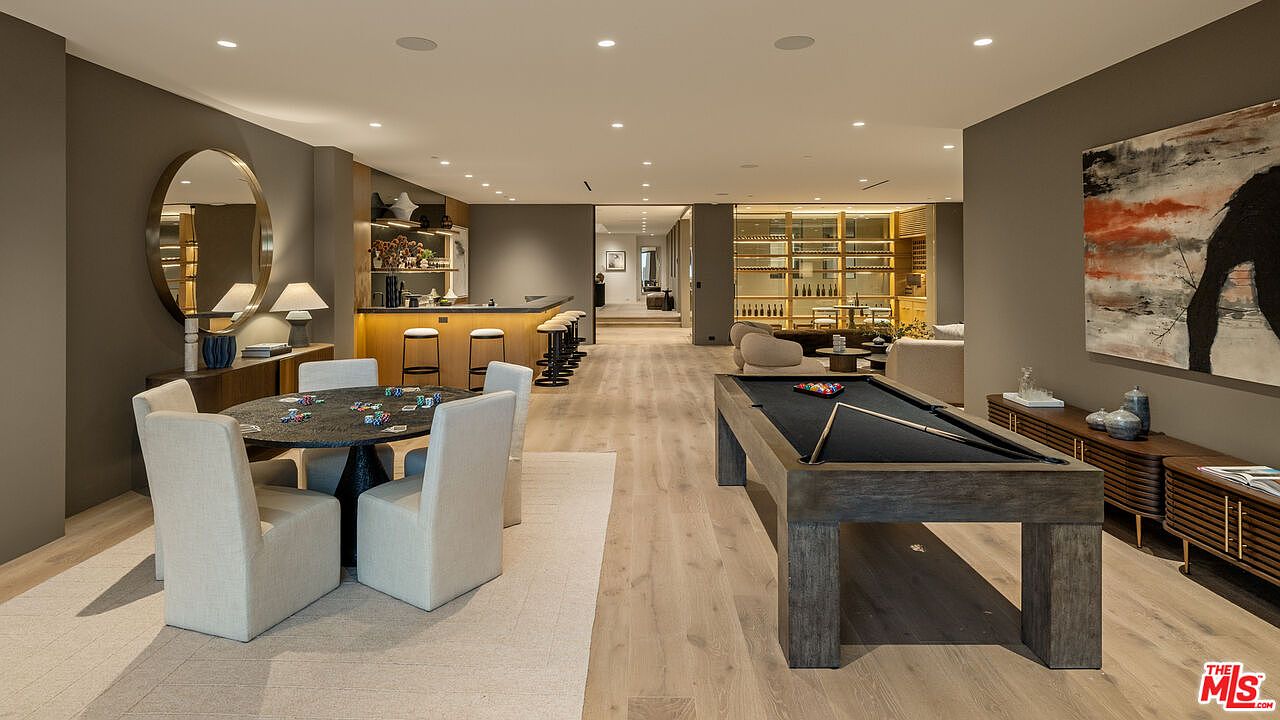 This expansive, open-concept entertainment space features a sophisticated game area with a pool table in the foreground and a circular dining or card table set for four. In the background, a sleek bar with integrated lighting and bar stools creates a refined lounge atmosphere, while light wood flooring runs throughout the entire room. The space is decorated with contemporary art, a large circular mirror, and neutral-toned furniture, offering a luxurious and inviting environment for social gatherings.