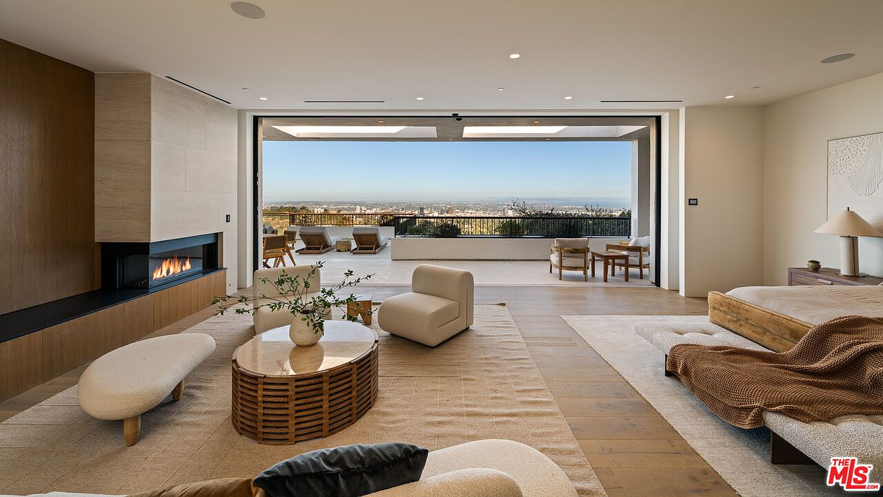 This expansive primary bedroom features a seamless transition to an outdoor terrace through wide-opening glass doors that frame a panoramic city view. The room is anchored by a neutral-toned rug, a low-profile bed, and a seating area centered around a linear fireplace with a tall stone-clad surround. The aesthetic is minimalist and sophisticated, utilizing light wood flooring and a soft, monochromatic color palette to emphasize the connection between the indoor living space and the horizon.