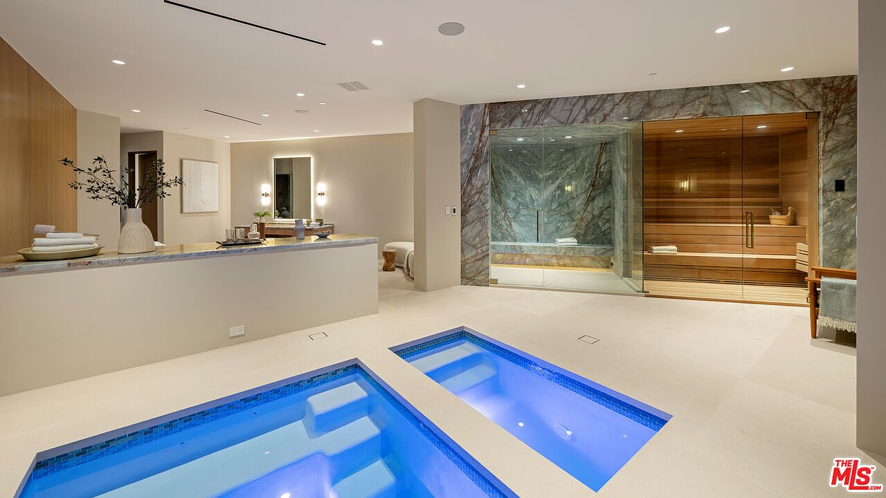 This luxurious spa-inspired bathroom features a sunken indoor pool with blue lighting, adjacent to a glass-enclosed sauna and a separate shower area with striking stone wall cladding. The space is designed with a neutral color palette, light-toned flooring, and a minimalist vanity area in the background. The perspective captures the serene, high-end wellness atmosphere of the room, emphasizing the seamless integration of relaxation amenities.