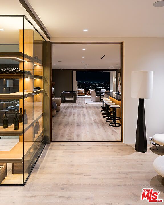 This image captures a sophisticated interior space featuring a glass-enclosed wine cellar on the left with illuminated shelving. Beyond the cellar, a wide doorway opens into a spacious lounge area with light wood flooring, a bar with stools, and a view of a city skyline at night. The perspective is from a hallway, highlighting the seamless transition between the modern storage area and the expansive entertainment space.