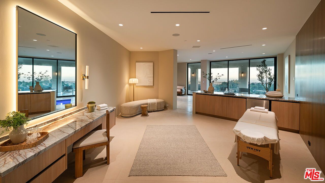 This luxurious spa or wellness room features a massage table, a vanity area with a large mirror, and a curved bench seat. The space is decorated in a neutral, calming palette with light-toned walls, stone surfaces, and warm wood accents. Large glass doors provide a seamless transition to an outdoor area, enhancing the serene and high-end atmosphere of the room.