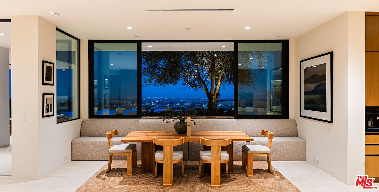 This elegant dining area features a light wood table paired with four matching chairs, set against a long, upholstered built-in bench. The space is framed by expansive, large windows that offer a stunning, panoramic view of a city skyline at dusk, accented by a mature tree just outside. The room is grounded by a textured area rug and decorated with framed artwork on the side walls, creating a sophisticated and serene atmosphere.
