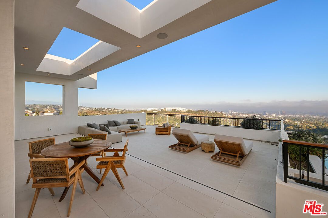 This expansive, modern balcony offers a panoramic view of the surrounding landscape and city skyline under a clear blue sky. The space is furnished with a circular wooden dining set, a comfortable lounge area with modular seating, and two wooden chaise lounges, all set against a clean, neutral-toned deck. A unique architectural feature includes a large, open-air cutout in the roof structure, allowing natural light to flood the partially covered outdoor living area.