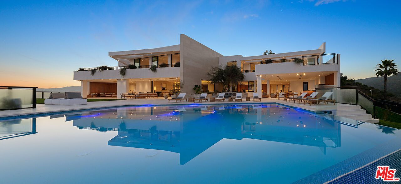 This expansive modern estate features a large, geometric infinity-edge pool that reflects the twilight sky and the home's architecture. The multi-level exterior showcases clean lines, light-toned stone cladding, and numerous lounge areas furnished with contemporary outdoor seating. The perspective is captured from the pool deck, emphasizing the seamless transition between the indoor living spaces and the outdoor entertainment area.
