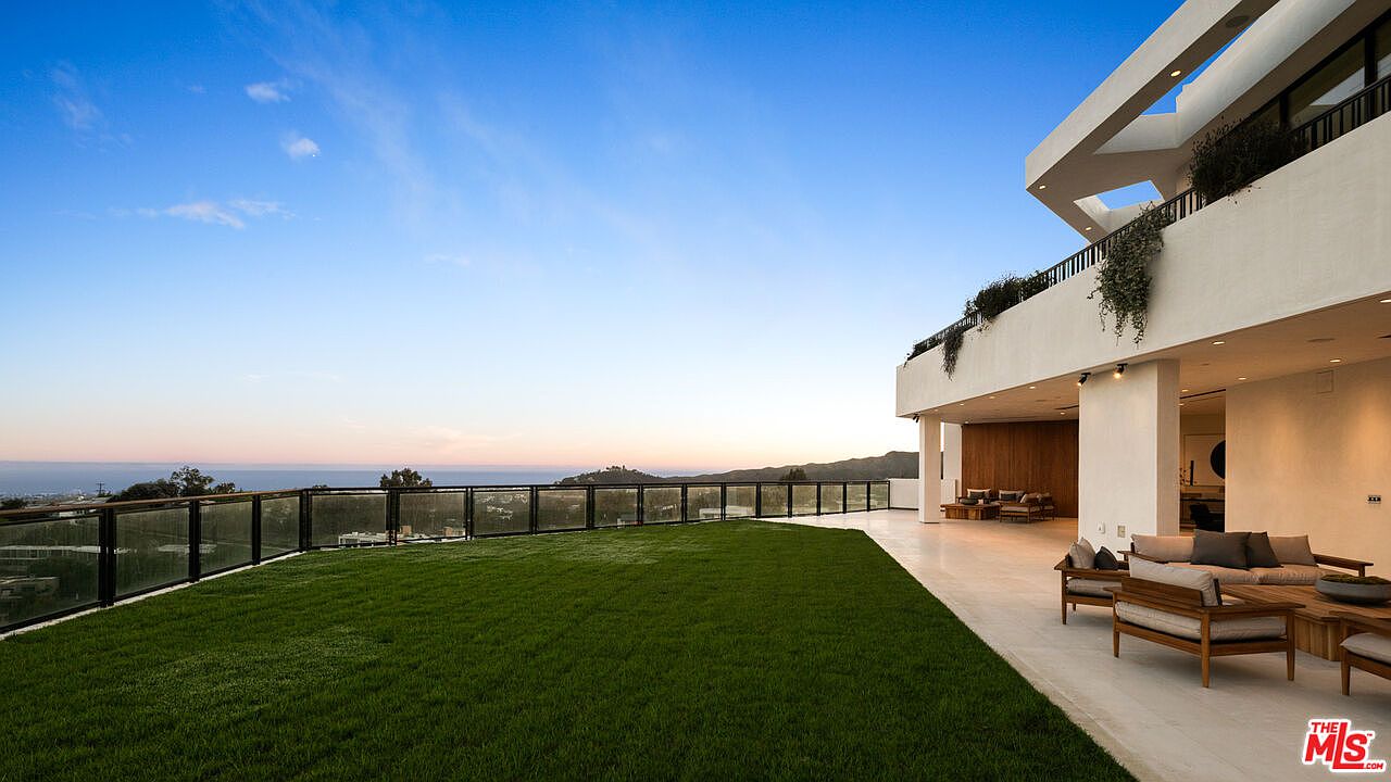 This expansive outdoor terrace features a manicured lawn bordered by a sleek glass railing that overlooks a scenic, distant landscape. A covered patio area provides a shaded lounge space furnished with comfortable seating, seamlessly connecting the indoor and outdoor living areas. The modern architectural design emphasizes clean lines and a bright, airy aesthetic, perfect for sunset entertaining.