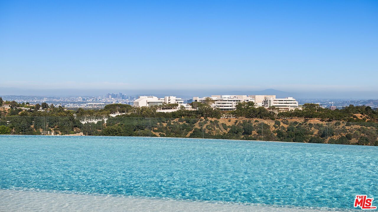 This image captures a stunning infinity-edge pool that seamlessly blends into a panoramic view of the Los Angeles skyline and the Getty Center. The foreground features the shimmering, turquoise water of the pool, while the background showcases a lush, rolling hillside leading to the distant urban landscape under a clear blue sky. The perspective is elevated, emphasizing the luxurious and serene atmosphere of the property's outdoor living space.
