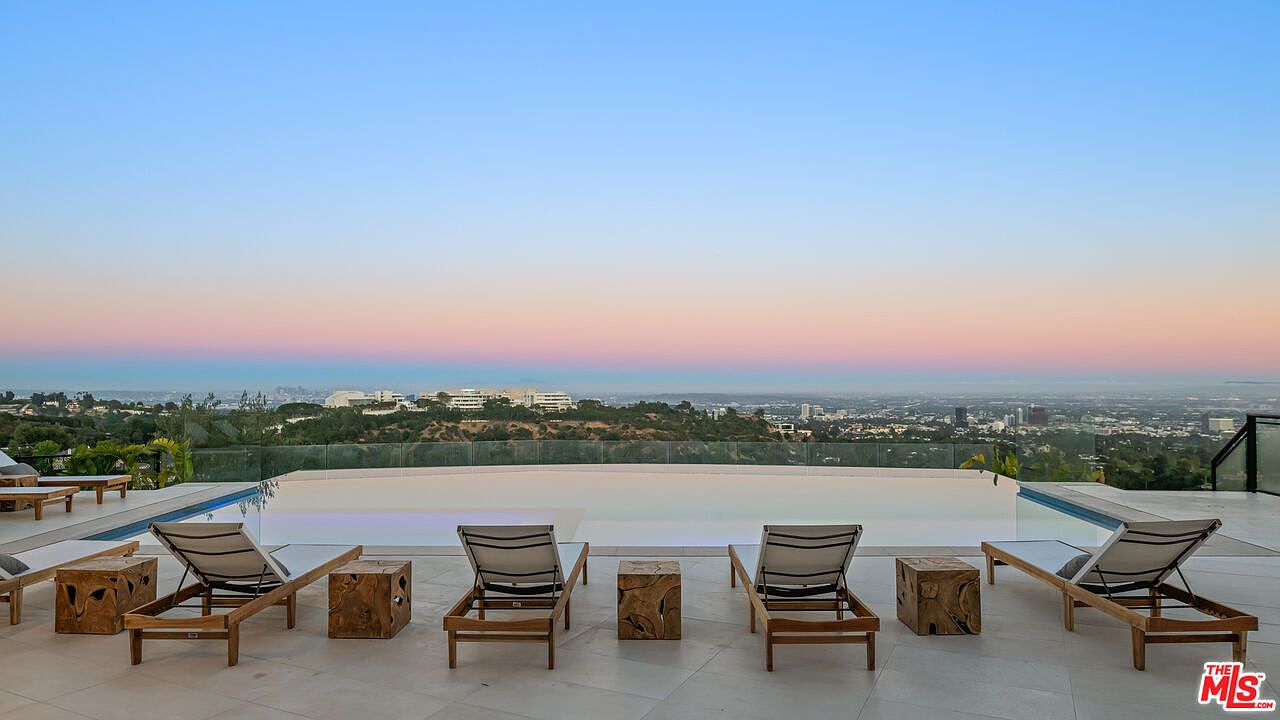 This image captures a luxurious infinity-edge pool overlooking a sprawling cityscape at dusk. The foreground features a sleek stone deck furnished with several modern lounge chairs and rustic wooden side tables, creating a serene outdoor retreat. The composition emphasizes the seamless transition between the pool's edge and the expansive horizon, highlighting the property's elevated vantage point.