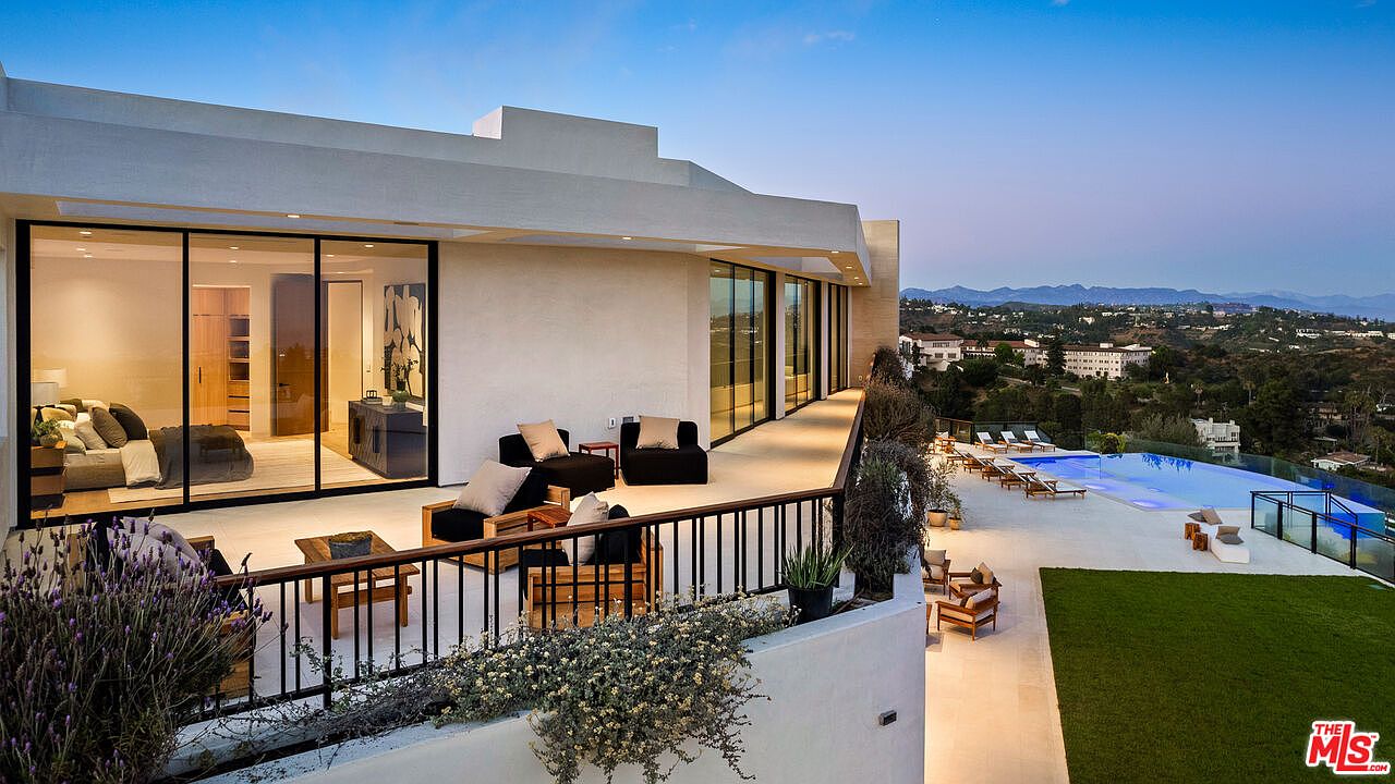 This elevated terrace offers a sweeping view of the surrounding hills and a lower-level pool area, blending indoor and outdoor living through expansive glass sliding doors. The space is furnished with modern outdoor seating, including armchairs and a coffee table, set against a backdrop of a minimalist, light-toned architectural structure. The scene is captured during the golden hour, highlighting the clean lines of the balcony railing and the serene, resort-like atmosphere of the property.