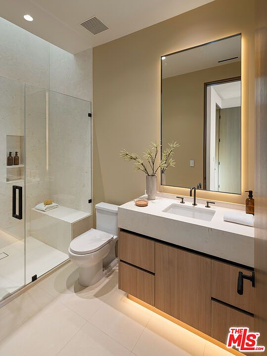 This modern bathroom features a sleek vanity with light wood cabinetry, a stone countertop, and a large, backlit mirror. To the left, a glass-enclosed shower includes a built-in bench and a recessed niche for toiletries, while a white toilet sits adjacent to the vanity. The space is finished with neutral-toned walls and light-colored flooring, creating a clean and contemporary aesthetic.
