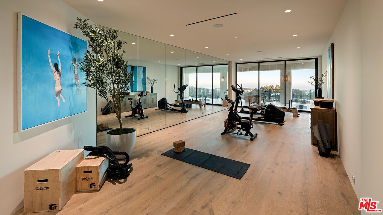 This spacious home gym features light wood flooring and a wall of mirrors that enhances the sense of depth and brightness. The room is equipped with various fitness gear, including a stationary bike, a rowing machine, and wooden plyometric boxes, while a large, vibrant artwork of swimmers adds a pop of color. Large sliding glass doors open to an outdoor area, allowing for natural light and a seamless transition to the exterior.
