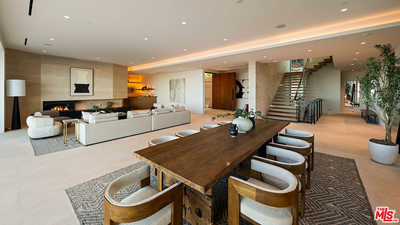 This expansive open-concept living and dining area features a neutral color palette with light stone flooring and warm wood accents. The living space is anchored by a fireplace with a tall stone-clad chimney breast, while the dining area showcases a large wooden table surrounded by modern chairs. The perspective captures the seamless flow between the seating area, the dining space, and the staircase leading to the upper level, creating a sophisticated and airy atmosphere.