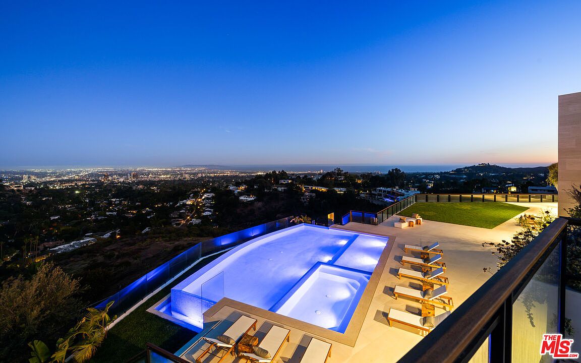 This high-angle, cinematic shot captures a luxurious infinity-edge pool illuminated in vibrant blue at twilight, overlooking a sprawling city skyline and the distant ocean. The expansive deck features multiple lounge chairs and a manicured lawn area, creating a sophisticated outdoor living space. The perspective emphasizes the dramatic elevation and the seamless integration between the modern architecture and the panoramic horizon.