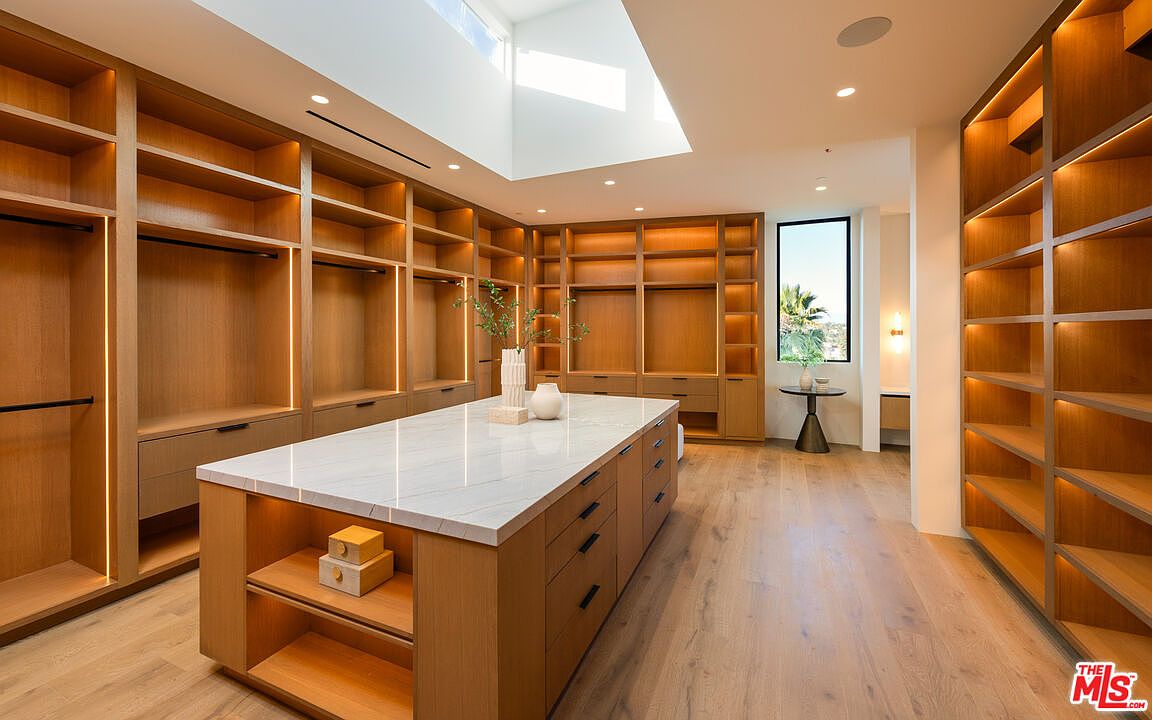This expansive walk-in closet features custom light wood cabinetry with integrated LED lighting throughout the shelving units. A large central island with a stone countertop provides additional storage and workspace, while a skylight above fills the room with natural light. The space is finished with light wood flooring, creating a bright, organized, and luxurious atmosphere.