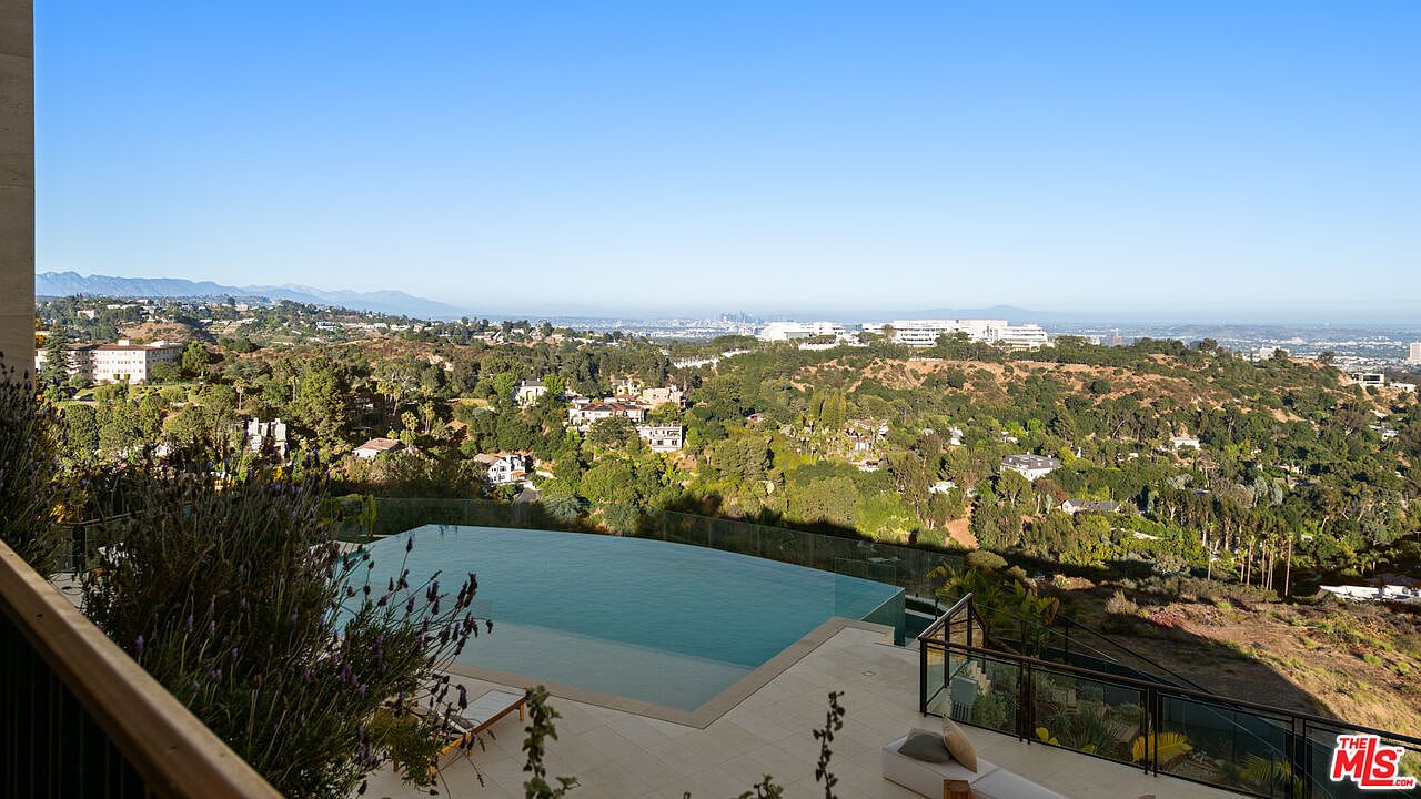 This elevated perspective captures a stunning infinity-edge pool overlooking a vast, lush hillside landscape and distant city skyline. The foreground features a sleek stone deck with lounge seating, providing a seamless transition between the private outdoor living space and the expansive natural scenery. The clear blue sky and panoramic views create a serene and luxurious atmosphere, highlighting the property's prime location.