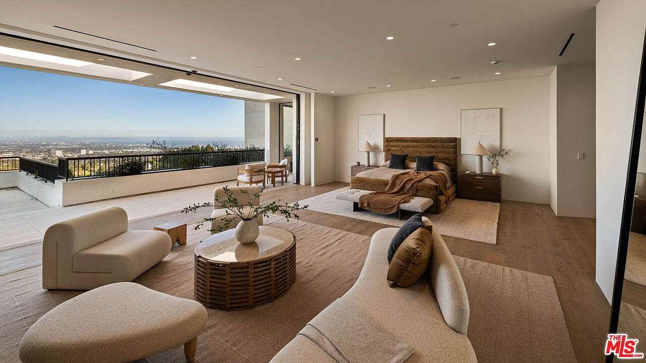 This expansive primary bedroom features a seamless transition to an outdoor balcony through large, retractable glass walls that offer panoramic city views. The room is furnished with a plush bed, a curved sofa, and a central seating area with a round coffee table, all set upon neutral-toned area rugs. The interior design is minimalist and sophisticated, utilizing a soft color palette and light wood flooring to create an airy, luxurious atmosphere.