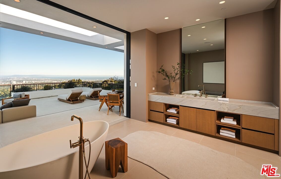 This luxurious primary bathroom features a freestanding soaking tub positioned to overlook a spacious outdoor terrace with panoramic city and ocean views. The interior includes a long vanity with a stone countertop, light wood cabinetry, and a large mirror, seamlessly blending indoor and outdoor living through expansive sliding glass doors. The aesthetic is modern and serene, emphasizing natural light and a neutral color palette.