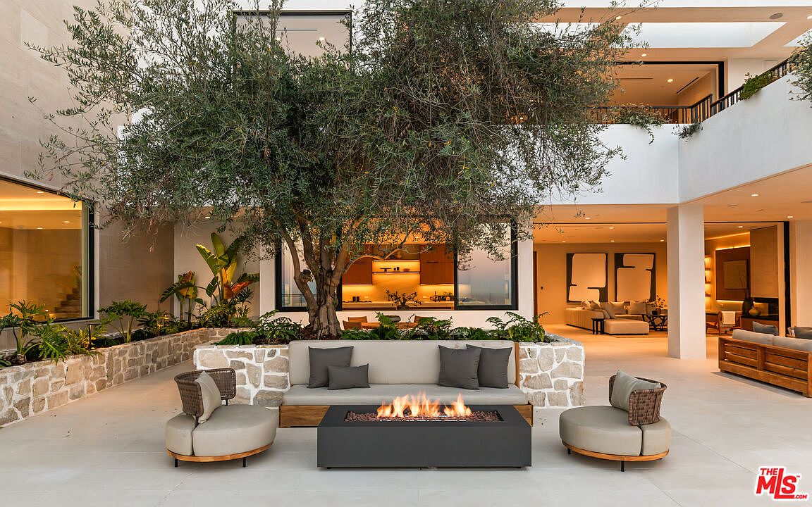 This luxurious outdoor patio features a central olive tree integrated into a stone-clad planter, serving as a focal point for the seating area. The space is furnished with a modern sofa and two circular lounge chairs arranged around a sleek, rectangular fire pit. The open-concept design seamlessly connects the outdoor lounge to the interior living spaces, creating a sophisticated and airy atmosphere perfect for entertaining.