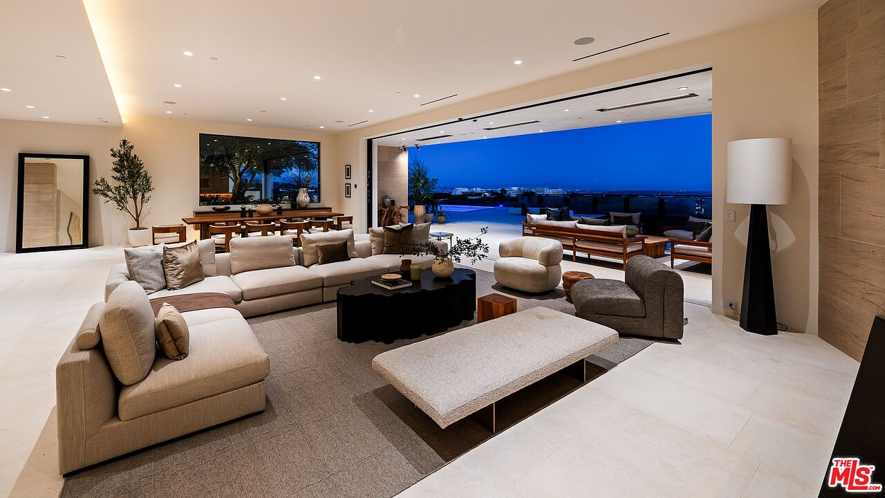 This expansive, open-concept living room features a neutral color palette and a large sectional sofa arranged around a unique, dark-toned coffee table. The space seamlessly transitions to an outdoor patio through a wide opening, highlighting a panoramic evening view of the city lights. A tall floor lamp stands near the corner, and the room is accented by a large mirror and a potted plant, creating a sophisticated and airy atmosphere.