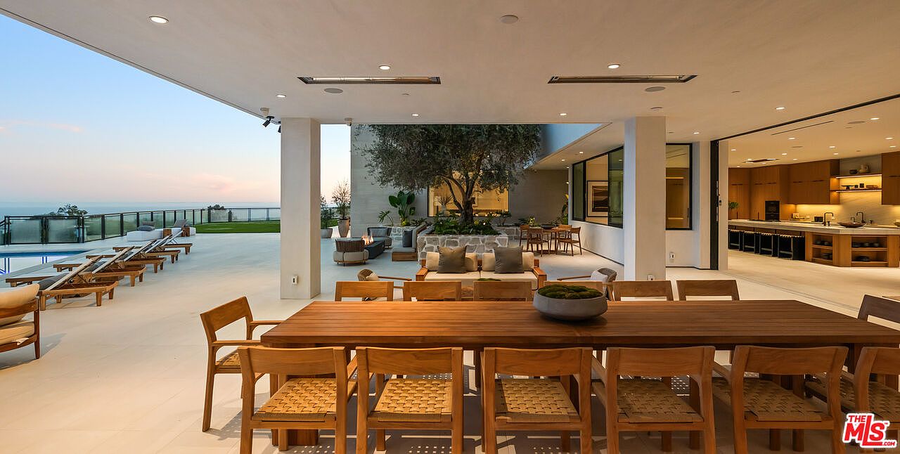 This expansive covered patio offers a seamless transition between indoor and outdoor living, featuring a long wooden dining table in the foreground and a lounge area with a fire pit beyond. The space is open to a pool deck with lounge chairs and a scenic ocean view, all sheltered by a modern structure with recessed lighting. A mature tree serves as a focal point in the central courtyard, enhancing the sophisticated and airy atmosphere of this luxury residence.