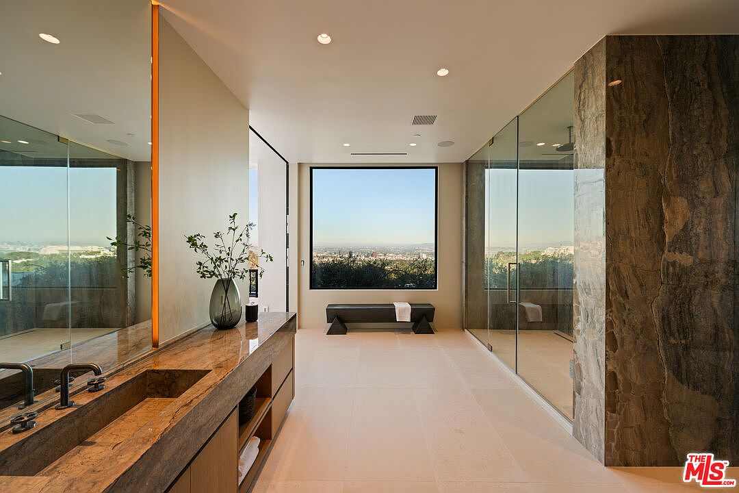 This luxurious primary bathroom features a long, stone-topped vanity with a built-in sink and a large mirror that reflects the room's clean, modern aesthetic. A glass-enclosed shower area sits to the right, while a minimalist bench is positioned in front of a large window that frames a panoramic view of the city. The space is finished with light-toned stone flooring and recessed lighting, creating a serene and sophisticated atmosphere.