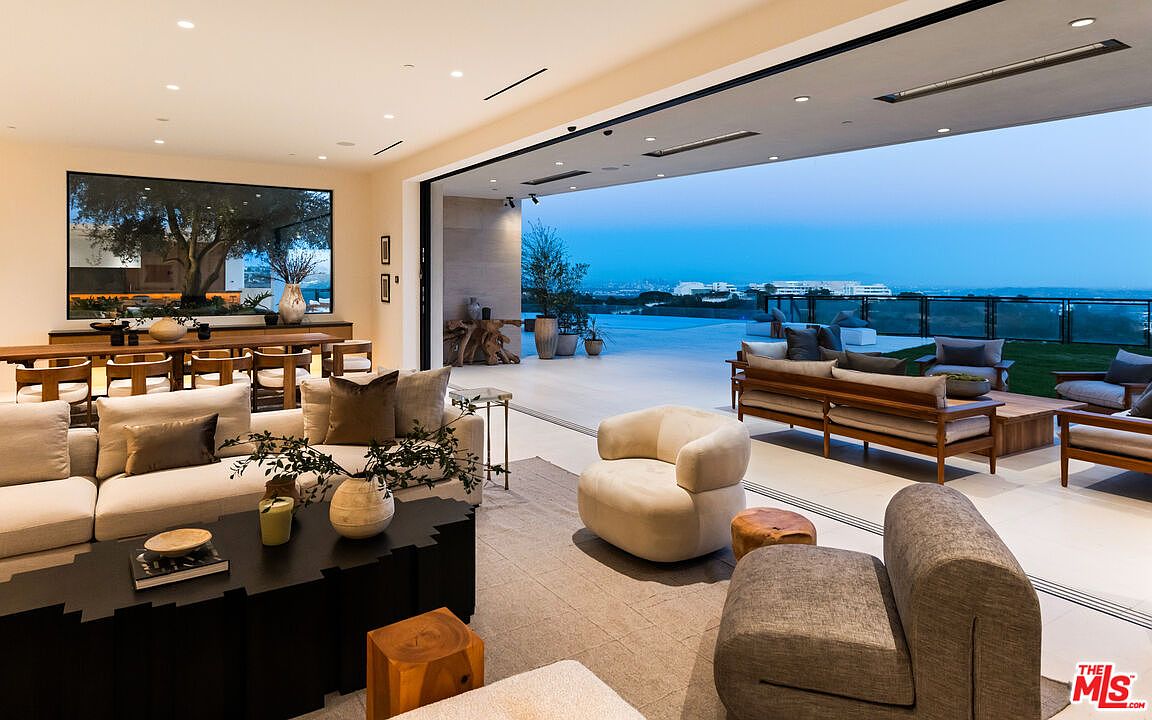 This expansive, open-concept living area seamlessly transitions to an outdoor patio through massive, retractable glass walls that offer panoramic views of the landscape. The interior features a neutral color palette with plush, contemporary seating, a dark textured coffee table, and a dining area visible in the background. The space is designed for luxury indoor-outdoor living, with recessed lighting and a sleek, modern aesthetic that emphasizes the connection to the horizon.