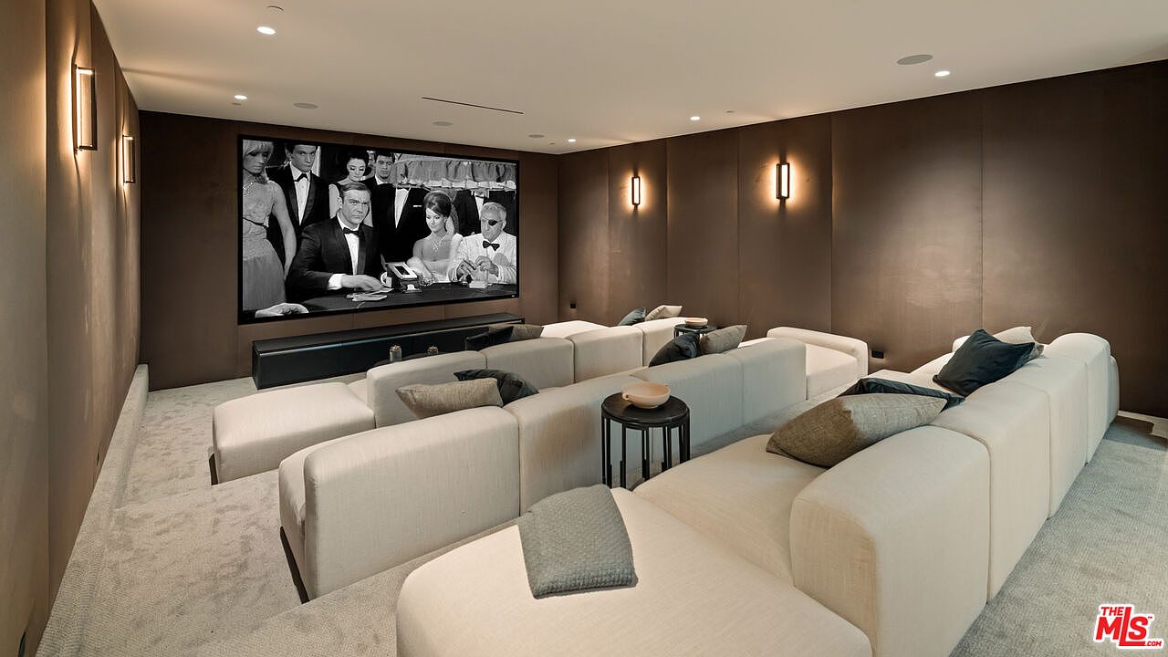 This image showcases a sophisticated home theater room featuring tiered seating with plush, light-colored modular sofas. The room is finished with dark, paneled walls and warm wall-mounted sconce lighting, creating an intimate and cinematic atmosphere. A large projection screen displays a classic film, serving as the focal point of this dedicated entertainment space.
