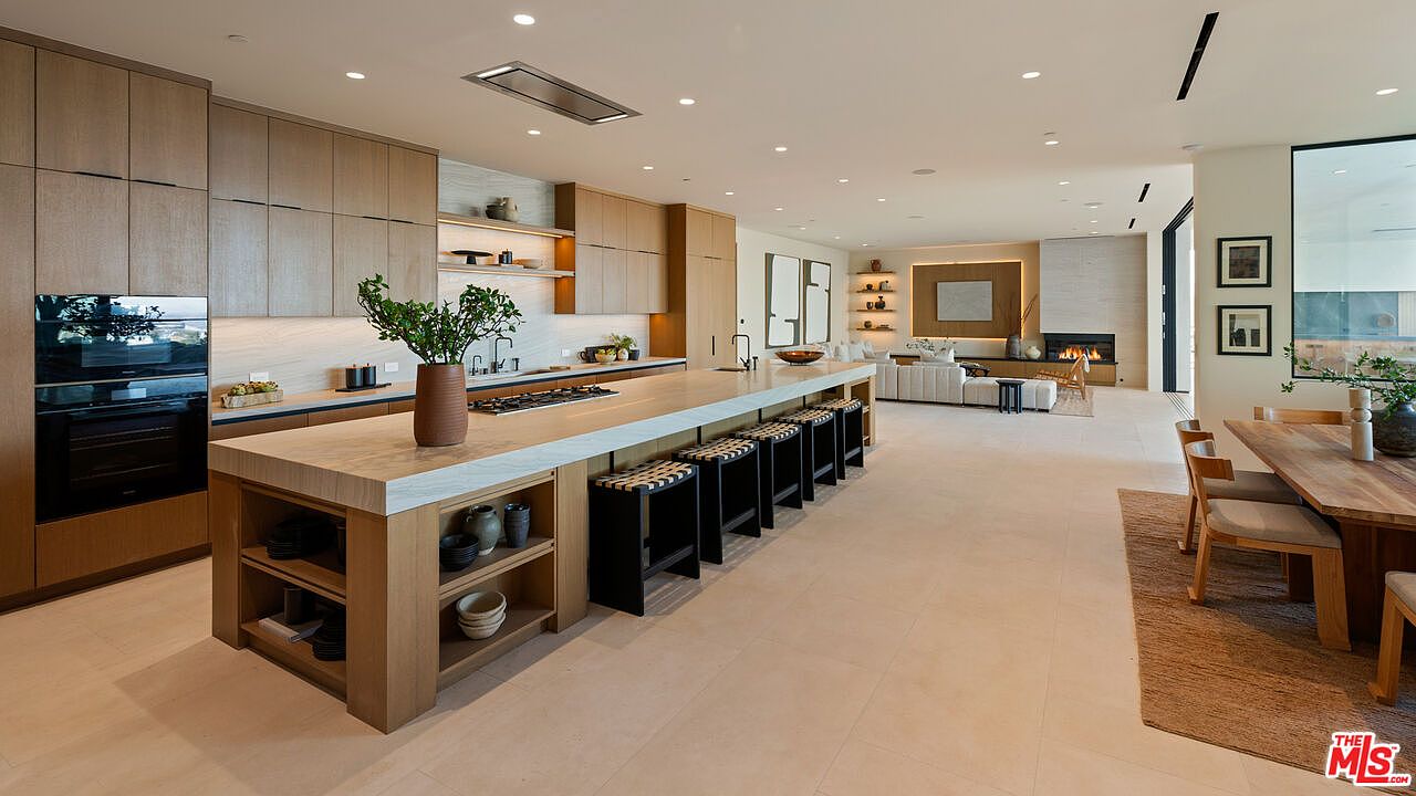 This expansive, open-concept kitchen features a massive central island with a light-toned stone countertop and integrated seating for five. The space is defined by sleek, light wood cabinetry, high-end built-in appliances, and a minimalist aesthetic that flows seamlessly into the adjacent living area. The perspective captures the depth of the home, highlighting the clean lines, recessed lighting, and the warm, inviting atmosphere created by the neutral color palette.