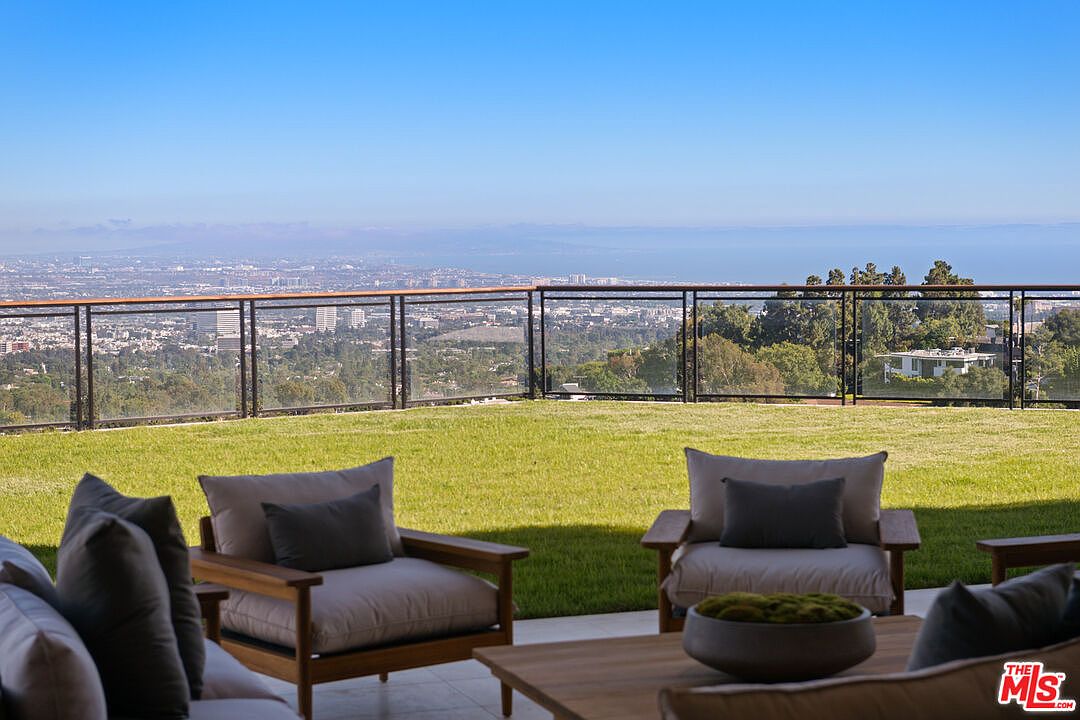 This elevated patio space features comfortable outdoor lounge furniture, including armchairs and a coffee table, set against a backdrop of a sprawling lawn. A sleek metal and glass railing provides an unobstructed, panoramic view of the city and coastline below. The perspective is from a seated position, emphasizing the serene and luxurious atmosphere of this outdoor living area.