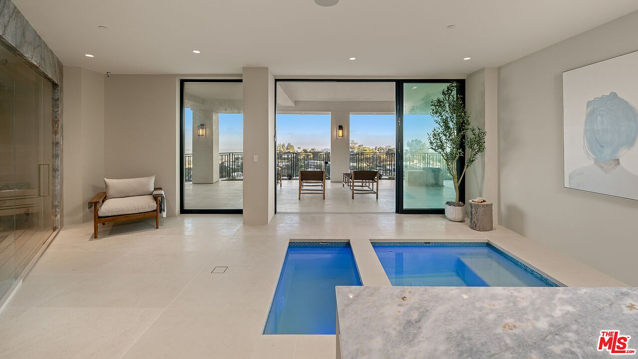 This bright, modern indoor spa area features a dual-section pool set into light-toned tile flooring. Large sliding glass doors open to an outdoor balcony, creating a seamless transition between the indoor space and the exterior view. To the left, a glass-enclosed sauna sits beside a comfortable lounge chair, while a piece of abstract art hangs on the wall to the right, contributing to the serene, minimalist aesthetic.