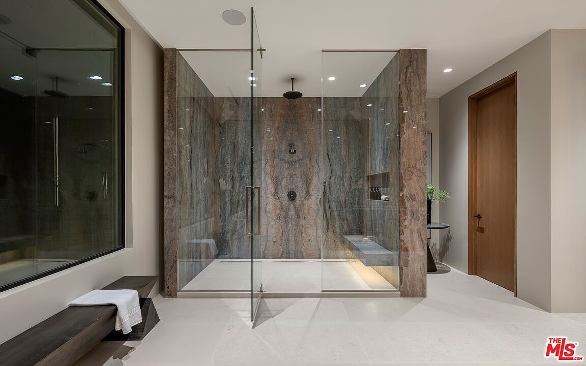 This luxurious primary bathroom features a large walk-in shower enclosed by glass panels, showcasing a striking, multi-toned stone wall with dramatic veining. A dark wood bench sits in the foreground, while a matching wooden door leads to an adjacent space, creating a sophisticated and cohesive aesthetic. The clean lines and neutral flooring emphasize the high-end, contemporary design of the room.