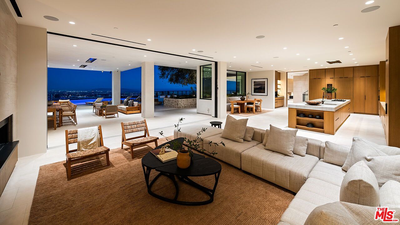 This expansive, open-concept living area seamlessly blends indoor and outdoor spaces through large sliding glass walls that open to a covered patio and pool deck. The room features a neutral color palette with a large sectional sofa, a textured area rug, and a modern coffee table, all centered around a fireplace with a tall stone surround. The adjacent kitchen area includes a substantial island with seating, showcasing a sophisticated, contemporary design with clean lines and warm wood cabinetry.