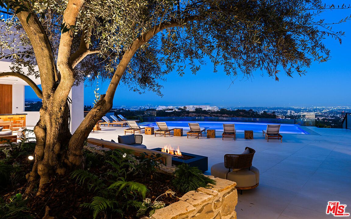 This luxurious outdoor terrace features a sleek infinity-edge pool overlooking a sprawling city skyline at twilight. The space is anchored by a large, mature olive tree and a modern linear fire feature, complemented by rows of lounge chairs on a light-toned stone deck. The perspective captures the seamless transition between the sophisticated patio living area and the expansive, illuminated urban horizon.
