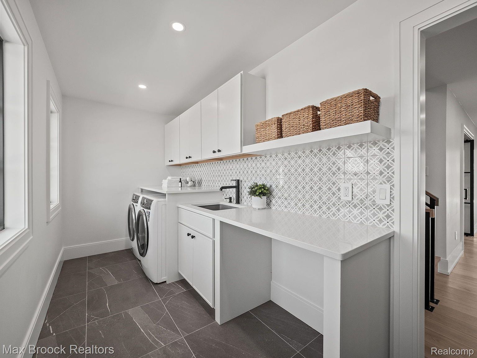 This is a bright and modern laundry room featuring white cabinetry, a countertop with a sink, and a washer and dryer set. The backsplash has a geometric pattern, and there are woven baskets on a shelf for storage. The flooring is a dark tile, and the room has a clean and organized appearance.