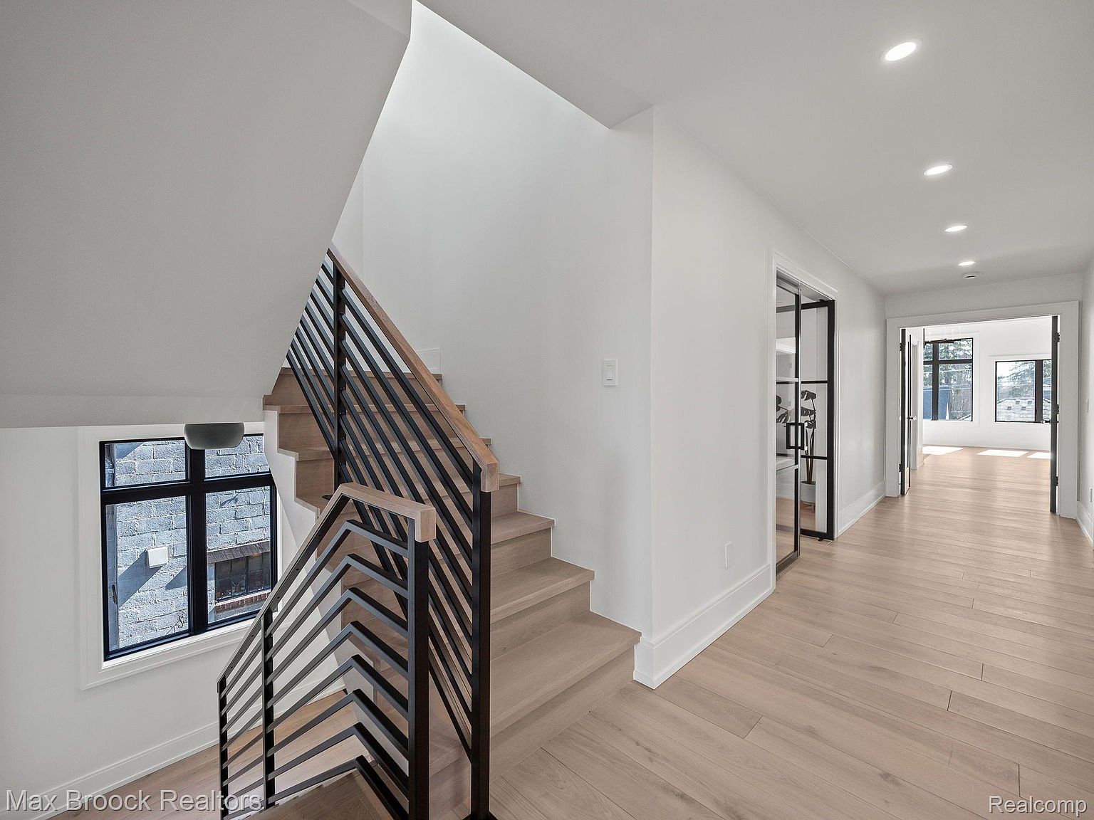 This interior shot showcases a modern hallway with wooden stairs and a sleek black railing. The light hardwood flooring and white walls create a bright and airy feel, complemented by recessed lighting. A glass-paneled door leads to another room, enhancing the open and contemporary design.